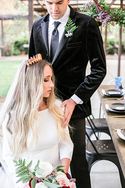  bride in a white lace gown and long sleeves and the groom in a black velvet tuxedo and a black long tie sitting together at the sweetheart table together close up