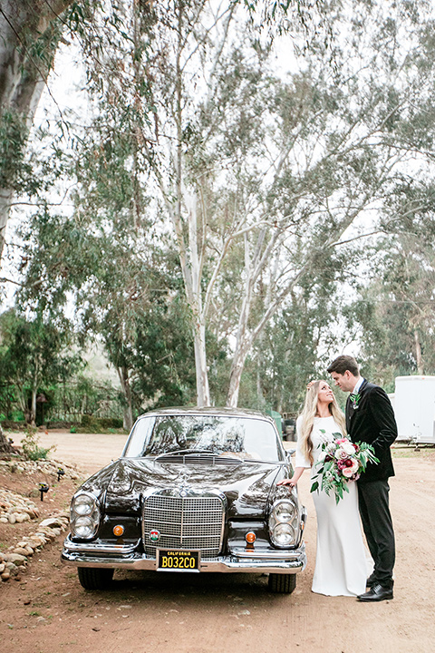 bride in a white lace gown and long sleeves and the groom in a black velvet tuxedo and a black long tie by a vintage car