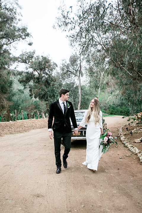  bride in a white lace gown and long sleeves and the groom in a black velvet tuxedo and a black long tie walking down the street together