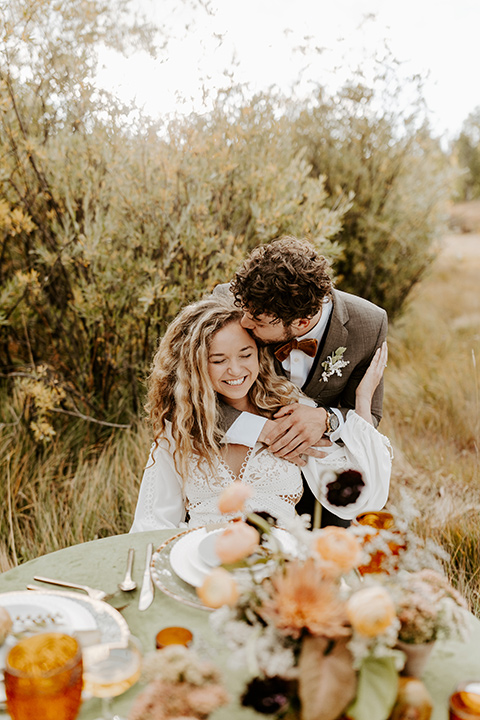  bride in a white lace gown with long flowing sleeves and high neckline with the groom in a café brown suit coat and blue pants with a gold velvet bow tie, sitting at a table with green linens and gold style flatware