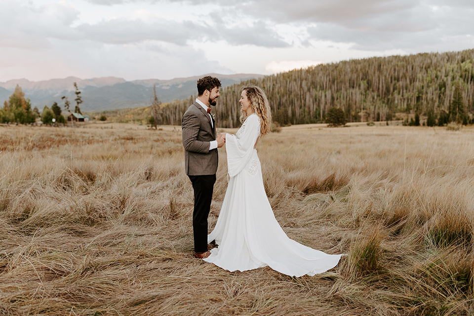  bride in a white lace gown with long flowing sleeves and high neckline with the groom in a café brown suit coat and blue pants with a gold velvet bow tie, exchanging vows in the fields