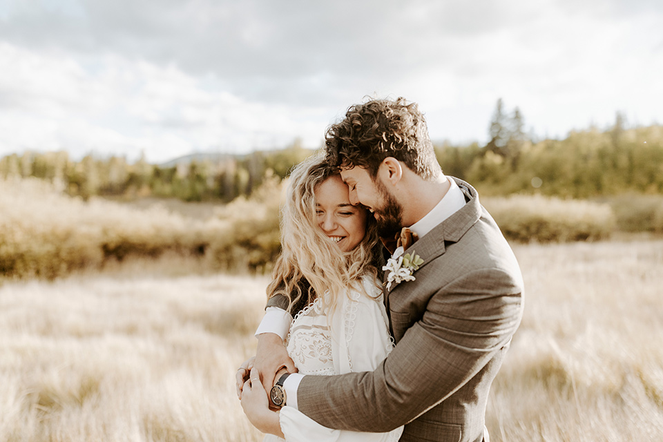  bride in a white lace gown with long flowing sleeves and high neckline with the groom in a café brown suit coat and blue pants with a gold velvet bow tie in an embrace