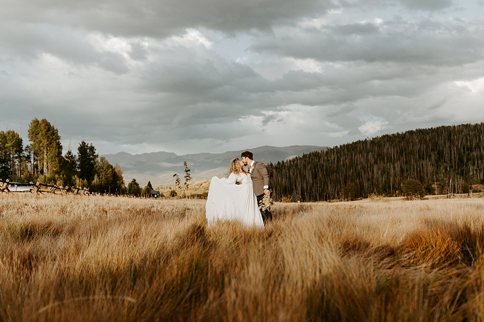  bride in a white lace gown with long flowing sleeves and high neckline with the groom in a café brown suit coat and blue pants with a gold velvet bow tie in an embrace in the fields