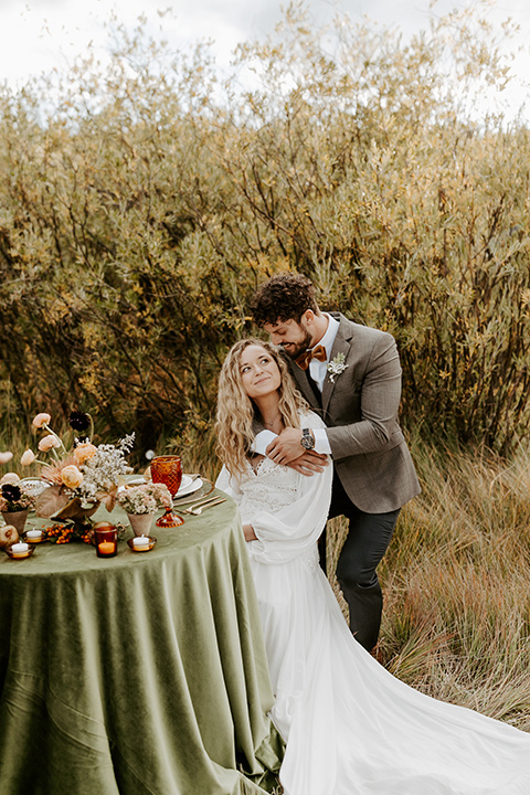  bride in a white lace gown with long flowing sleeves and high neckline with the groom in a café brown suit coat and blue pants with a gold velvet bow tie, sitting at a table with green linens and gold style flatware