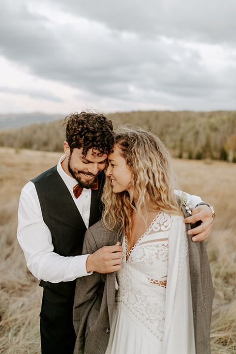  bride in a white lace gown with long flowing sleeves and high neckline with the groom in a café brown suit coat and blue pants with a gold velvet bow tie, with the groom giving up his jacket to his bride