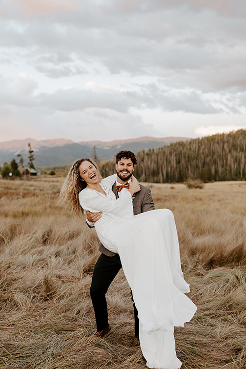  bride in a white lace gown with long flowing sleeves and high neckline with the groom in a café brown suit coat and blue pants with a gold velvet bow tie, groom spinning bride in his arms