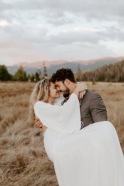  bride in a white lace gown with long flowing sleeves and high neckline with the groom in a café brown suit coat and blue pants with a gold velvet bow tie, with the groom holding up the bride