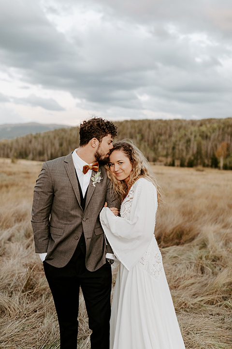  bride in a white lace gown with long flowing sleeves and high neckline with the groom in a café brown suit coat and blue pants with a gold velvet bow tie, staring at the camera