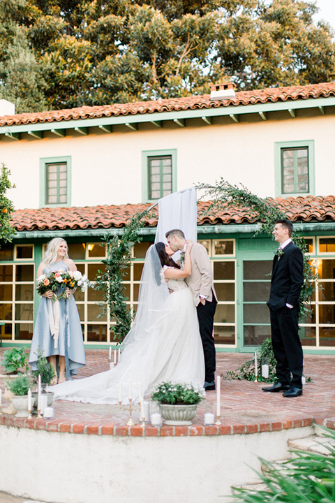  bride in a ballgown with a lace illusion neckline and long cathedral veil and the groom in a tan suit coat with black pants walking away 