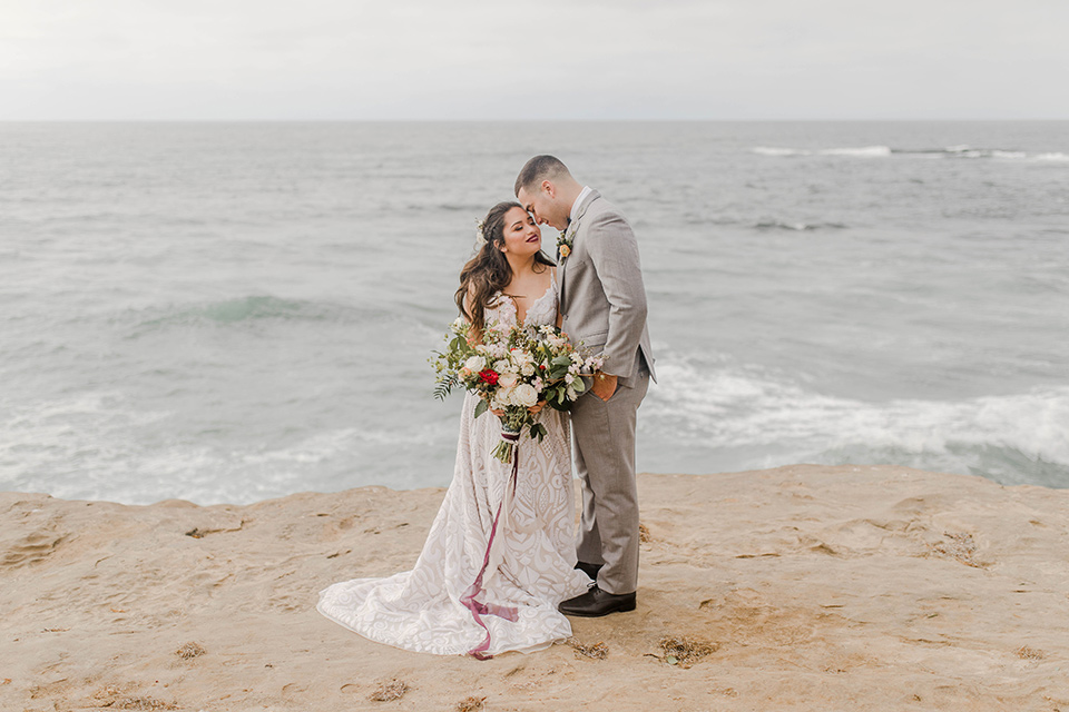  bride in a white gown with a flowing white skirt and low cut back and the groom in a light grey suit and charcoal bow tie near the beach