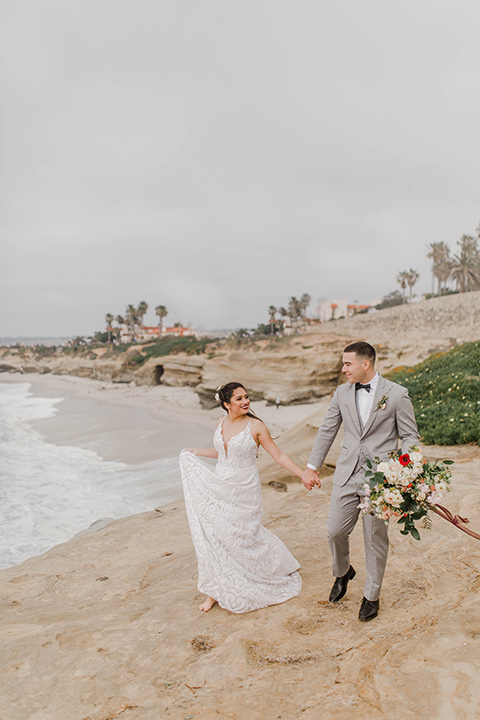  bride in a white flowing gown and a low cut back and the groom in a light grey suit with a white shirt and dark grey bow tie walking along the beach