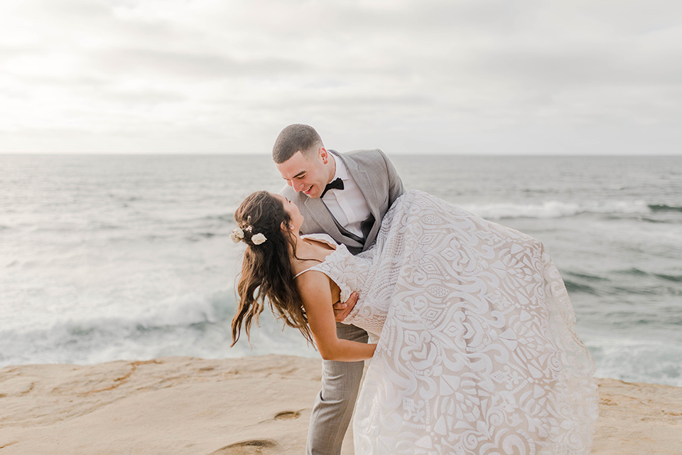  bride in a white gown with a flowing white skirt and low cut back and the groom in a light grey suit and charcoal bow tie near the beach