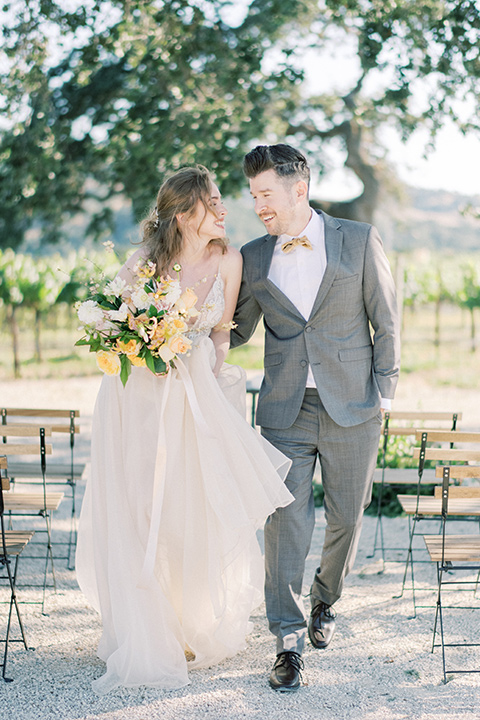  bride looking in a white flowing gown and the groom in a grey notch lapel suit with a gold velvet bow tie 