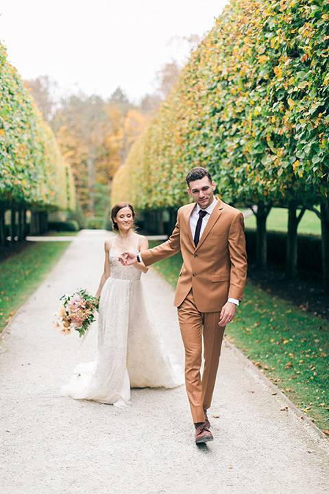  bride in a tulle ivory ballgown with a natural waist and jeweled detailing, the groom is in a caramel rust colored suit with a dark brown long tie, the walk down a path with trees and grass