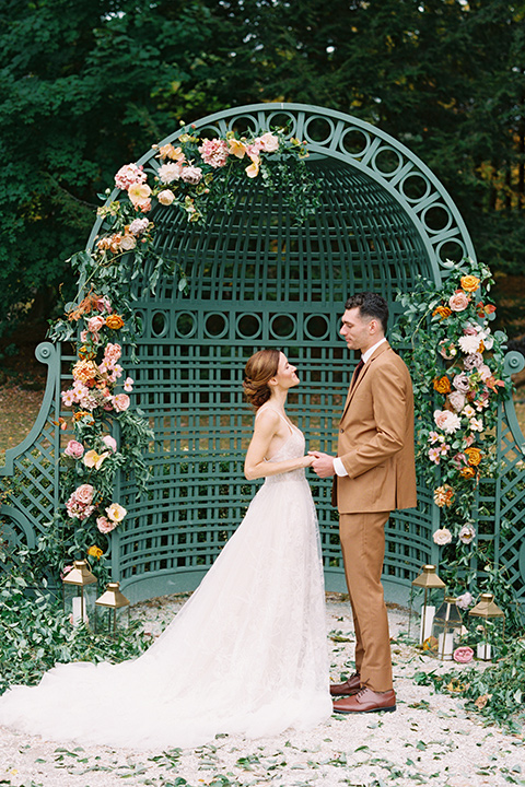  bride in a tulle ivory ballgown with a natural waist and jeweled detailing, the groom is in a caramel rust colored suit with a dark brown long tie, at the ceremony with green orange and pink floral arrangements