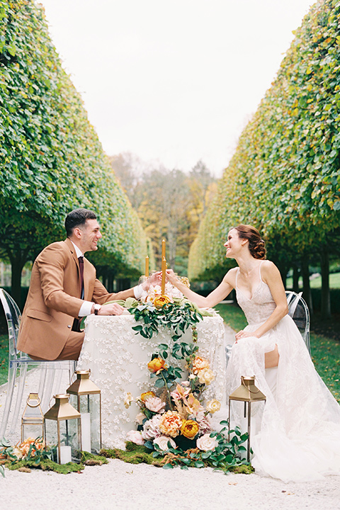  bride in a tulle ivory ballgown with a natural waist and jeweled detailing, the groom is in a caramel rust colored suit with a dark brown long tie, sitting at the table at the receotion