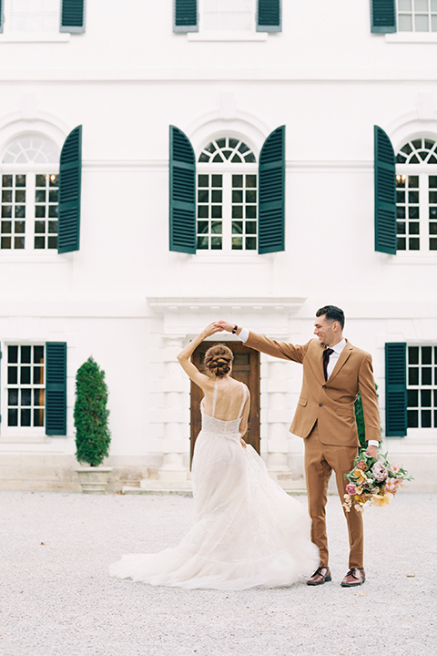  bride in a tulle ivory ballgown with a natural waist and jeweled detailing, the groom is in a caramel rust colored suit with a dark brown long tie dancing in front of the venue