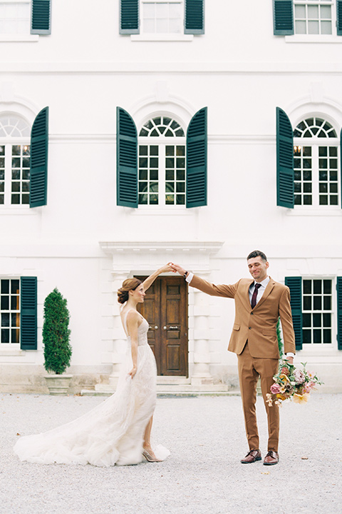  bride in a tulle ivory ballgown with a natural waist and jeweled detailing, the groom is in a caramel rust colored suit with a dark brown long tie, twirling around in front of the venue