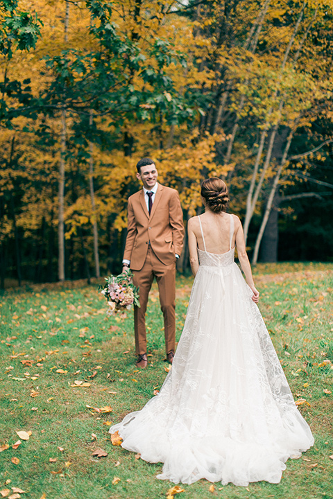  bride in a tulle ivory ballgown with a natural waist and jeweled detailing, the groom is in a caramel rust colored suit with a dark brown long tie they stand in a field together