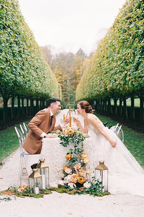  bride in a tulle ivory ballgown with a natural waist and jeweled detailing, the groom is in a caramel rust colored suit with a dark brown long tie kissing at the reception table