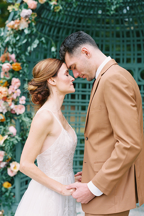  bride in a tulle ivory ballgown with a natural waist and jeweled detailing, the groom is in a caramel rust colored suit with a dark brown long tie touching their heads and smiling at the ceremony