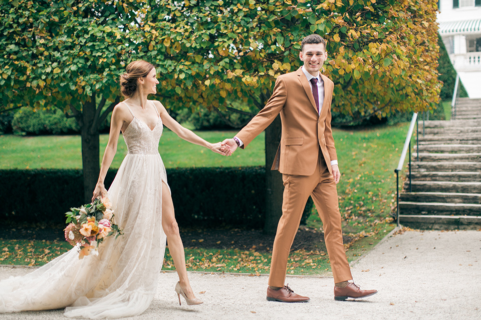  bride in a tulle ivory ballgown with a natural waist and jeweled detailing, the groom is in a caramel rust colored suit with a dark brown long tie, the walk down a path with trees and grass 