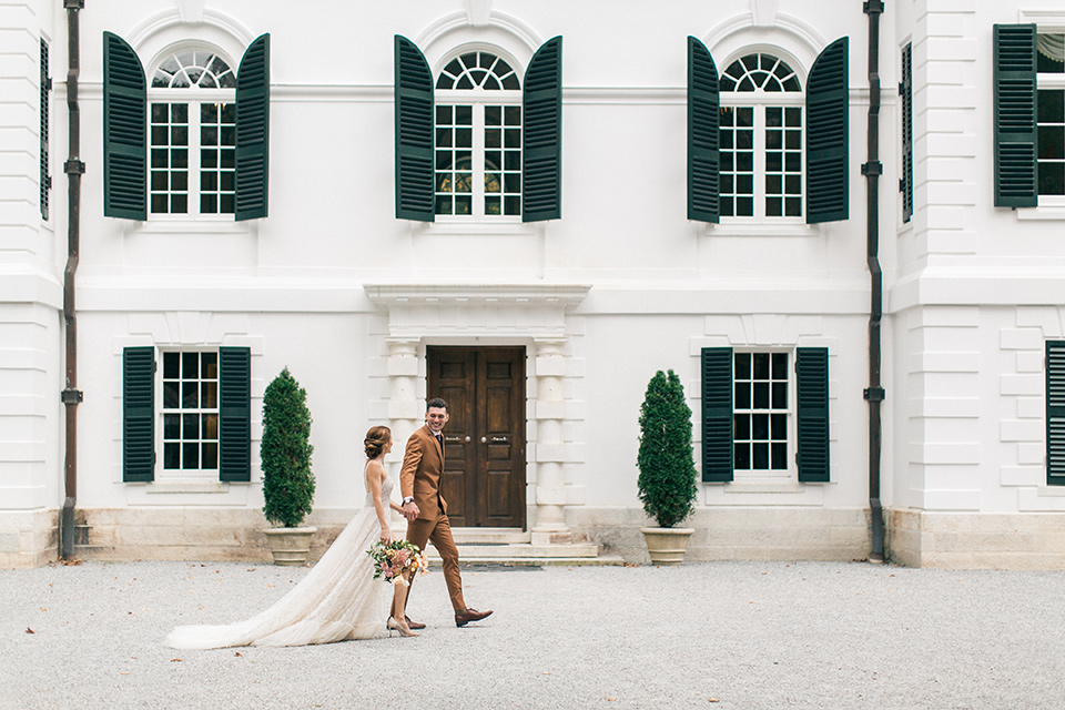  bride in a tulle ivory ballgown with a natural waist and jeweled detailing, the groom is in a caramel rust colored suit with a dark brown long tie, walking in front of the venue together 