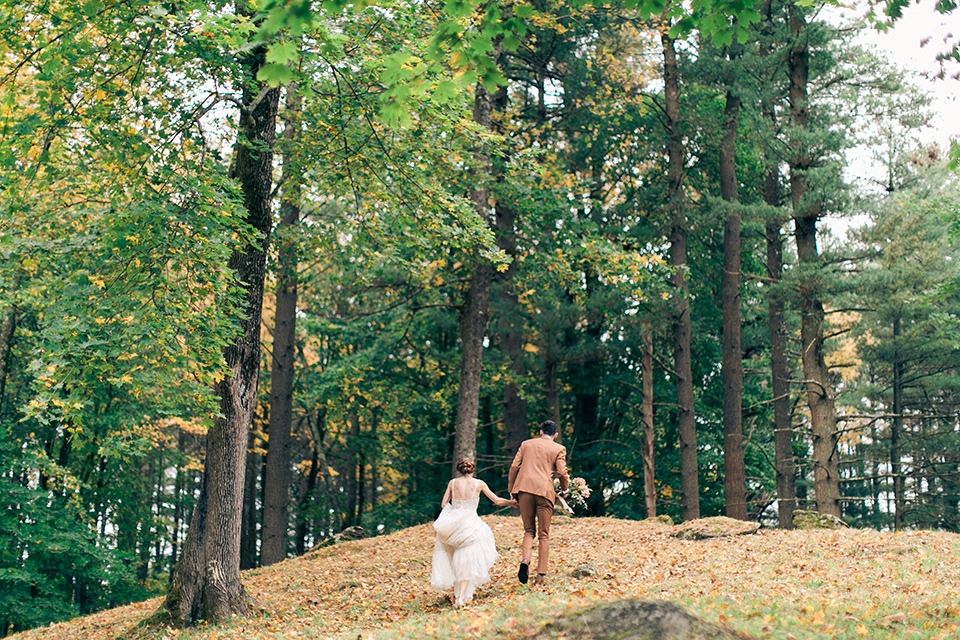  bride in a tulle ivory ballgown with a natural waist and jeweled detailing, the groom is in a caramel rust colored suit with a dark brown long tie, walking up a leaf covered hill on the venue property 