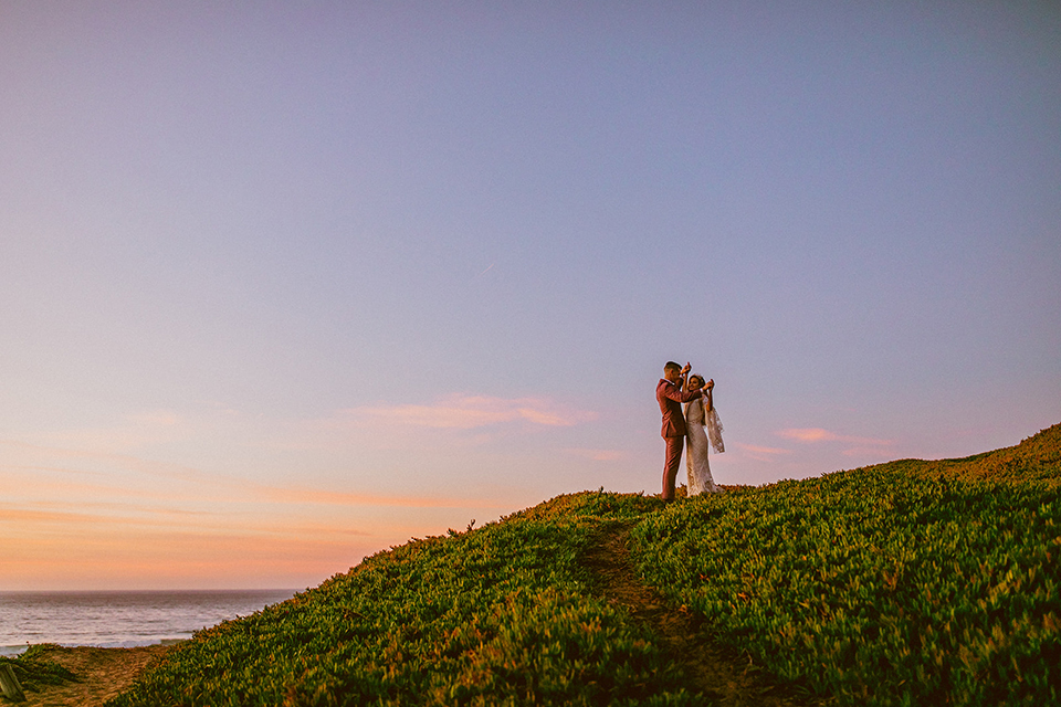  bride in a lace long sleeve gown with a plunging neckline and a boho headpiece and the groom in a rose pink suit with a gold velvet bow tie 