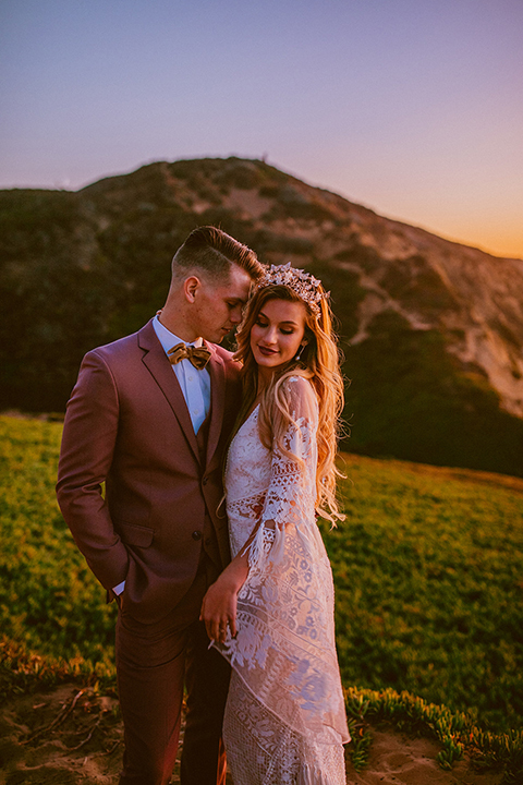  bride in a lace long sleeve gown with a plunging neckline and a boho headpiece and the groom in a rose pink suit with a gold velvet bow tie