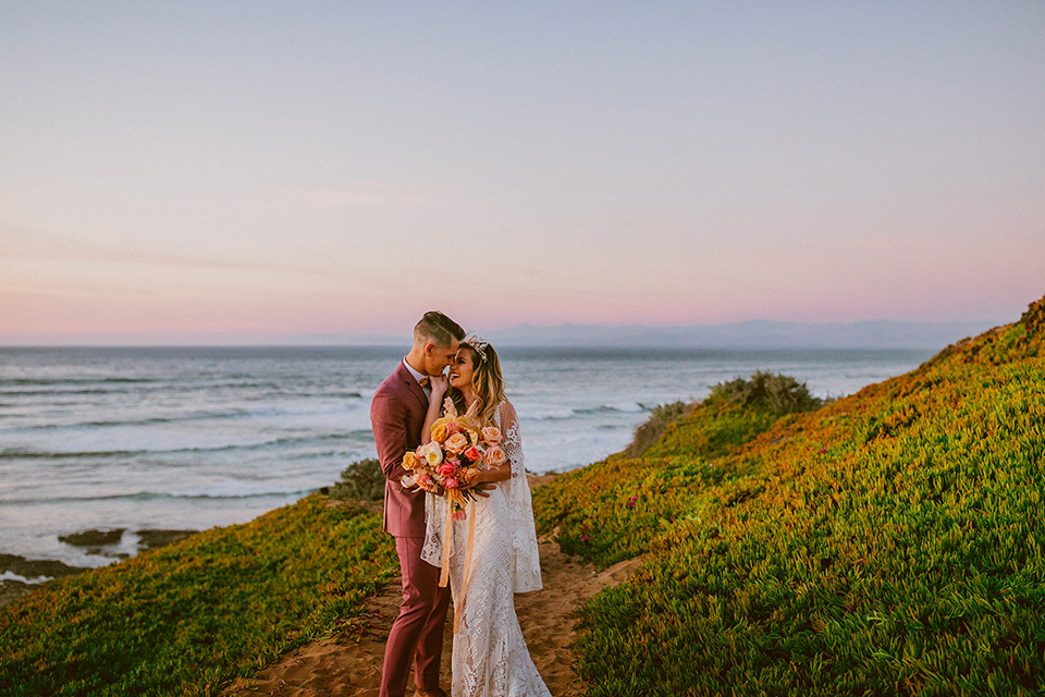  bride in a lace long sleeve gown with a plunging neckline and a boho headpiece and the groom in a rose pink suit with a gold velvet bow tie 