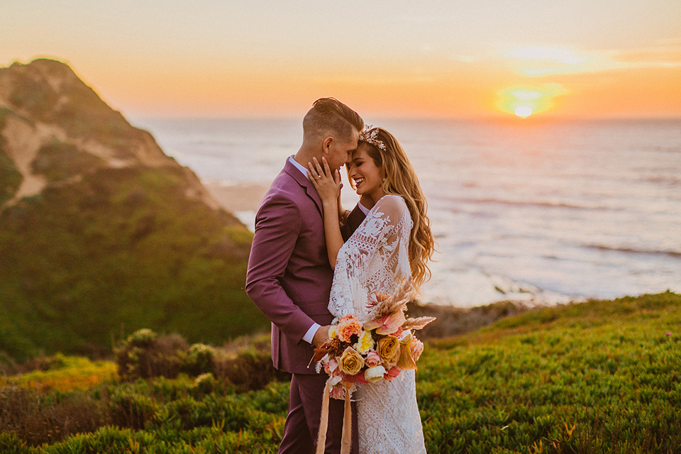  bride in a lace long sleeve gown with a plunging neckline and a boho headpiece and the groom in a rose pink suit with a gold velvet bow tie 