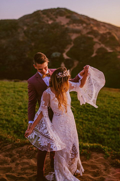  bride in a lace long sleeve gown with a plunging neckline and a boho headpiece and the groom in a rose pink suit with a gold velvet bow tie 