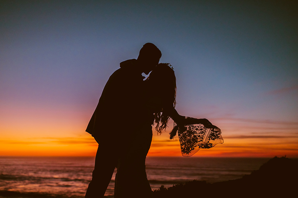  bride in a lace long sleeve gown with a plunging neckline and a boho headpiece and the groom in a rose pink suit with a gold velvet bow tie 