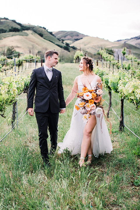 bride in a flowing bohemian gown with her hair in a loose braided bun and flowers and the groom in an asphalt grey suit