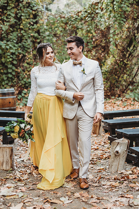   groom in an ivory paisley tuxedo and gold velvet bow tie and the bride in a two-piece gown with a yellow skirt and ivory long sleeve top 