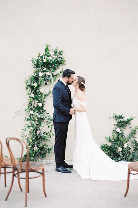  bride in a formfitting white lace gown with an off the shoulder detail  and the groom in a navy shawl lapel tuxedo