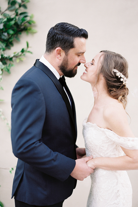  bride in a formfitting white lace gown with an off the shoulder detail  and the groom in a navy shawl lapel tuxedo 