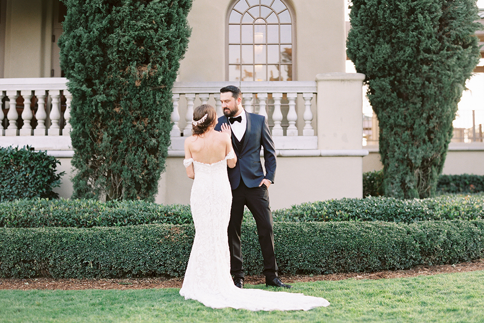  bride in a formfitting white lace gown with an off the shoulder detail  and the groom in a navy shawl lapel tuxedo 