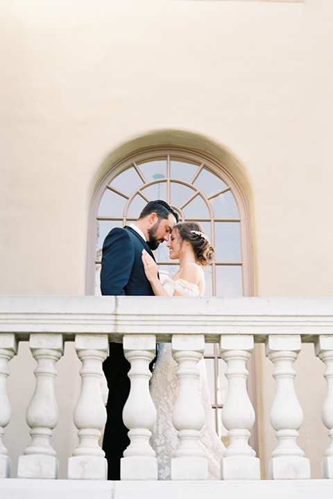  bride in a formfitting white lace gown with an off the shoulder detail  and the groom in a navy shawl lapel tuxedo