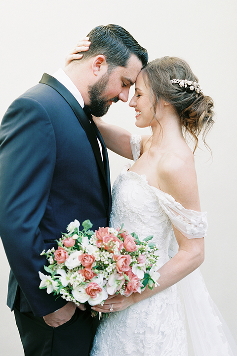  bride in a formfitting white lace gown with an off the shoulder detail  and the groom in a navy shawl lapel tuxedo