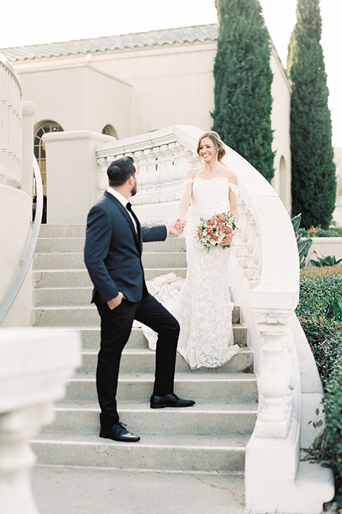  bride in a formfitting white lace gown with an off the shoulder detail  and the groom in a navy shawl lapel tuxedo 