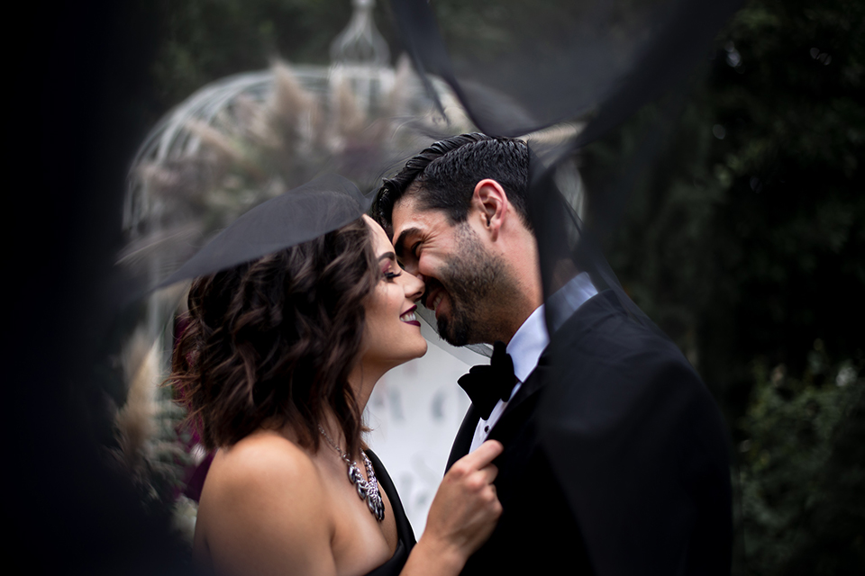  bride in a black gown with a black veil and the groom in a burgundy tuxedo with a black shirt and bow tie 