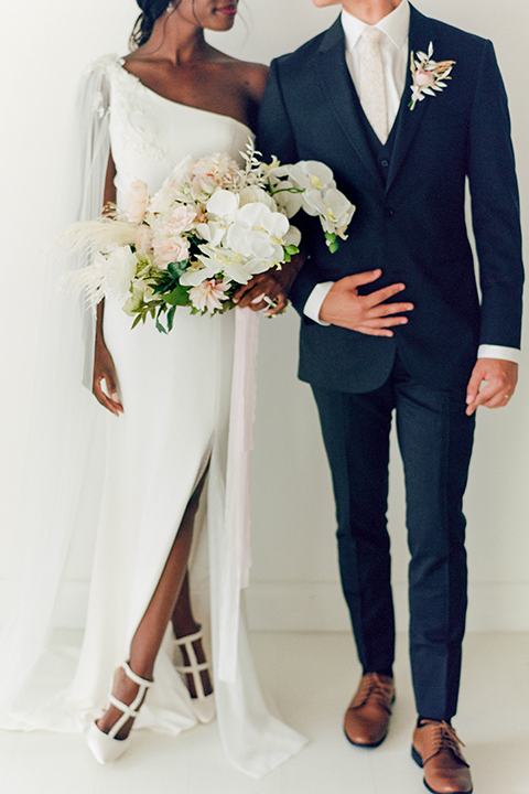  bride in a white formfitting modern gown and cape and the groom in a slate blue suit with a silver long tie 