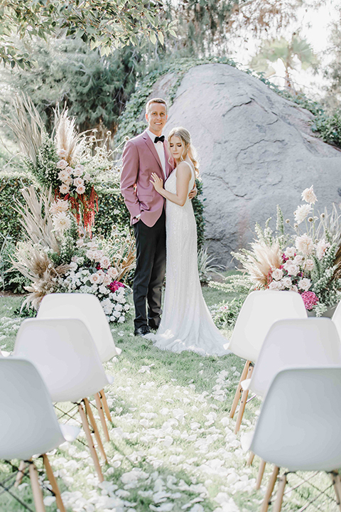  the bride and groom at the ceremony – the bride in a white lace gown and the groom in a rose pink coat and black pants