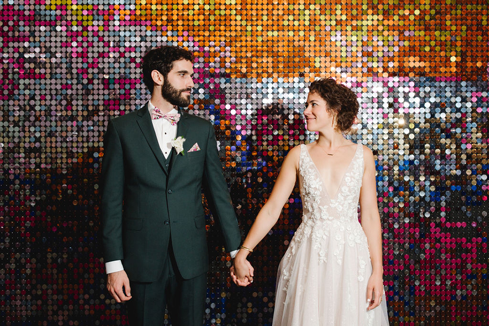  groom in a green suit with a pink floral bow tie and bride in a white a line gown with colorful flowers 