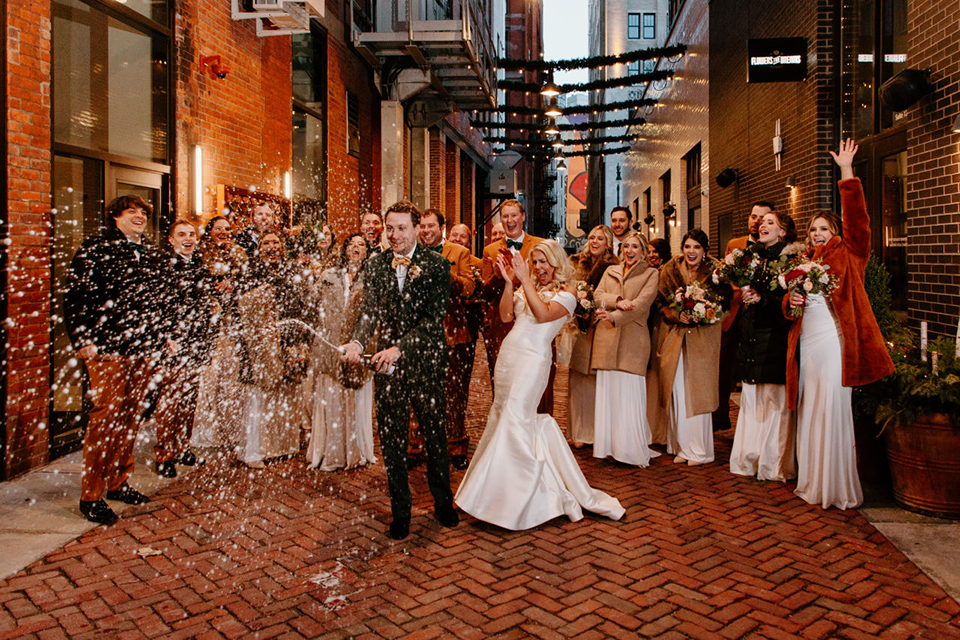  bride in a trumpet style gown and the groom in a green suit, the groomsmen in caramel notch lapel suits and the bridesmaids in ivory gowns 