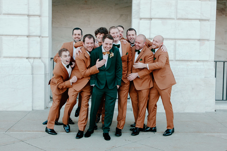  bride in a trumpet style gown and the groom in a green suit, the groomsmen in caramel notch lapel suits and the bridesmaids in ivory gowns 