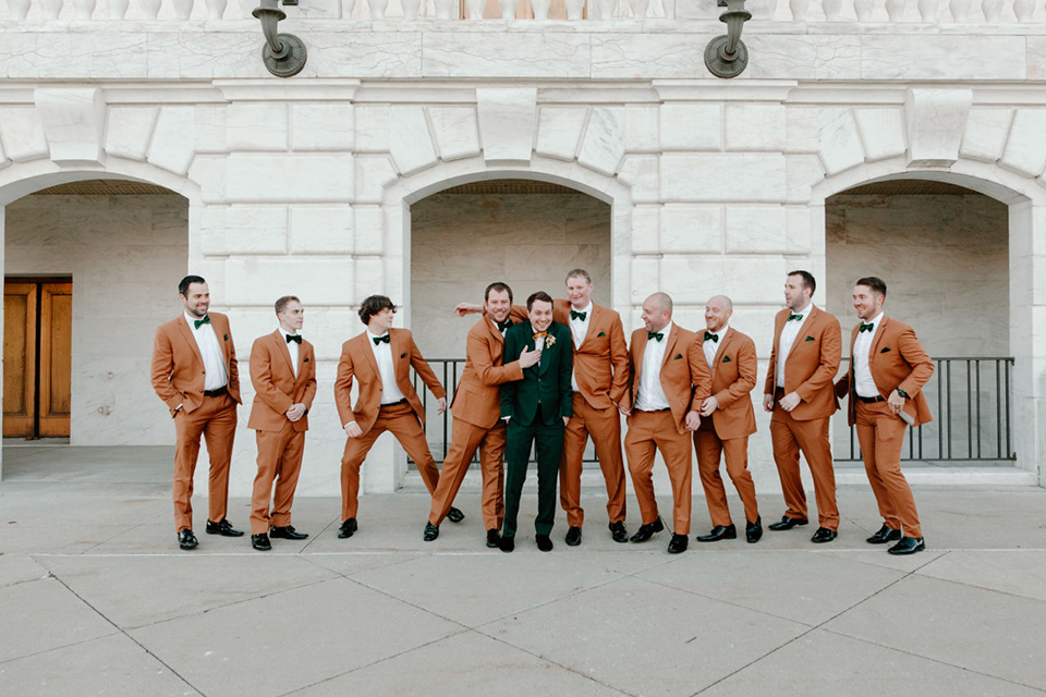  bride in a trumpet style gown and the groom in a green suit, the groomsmen in caramel notch lapel suits and the bridesmaids in ivory gowns 