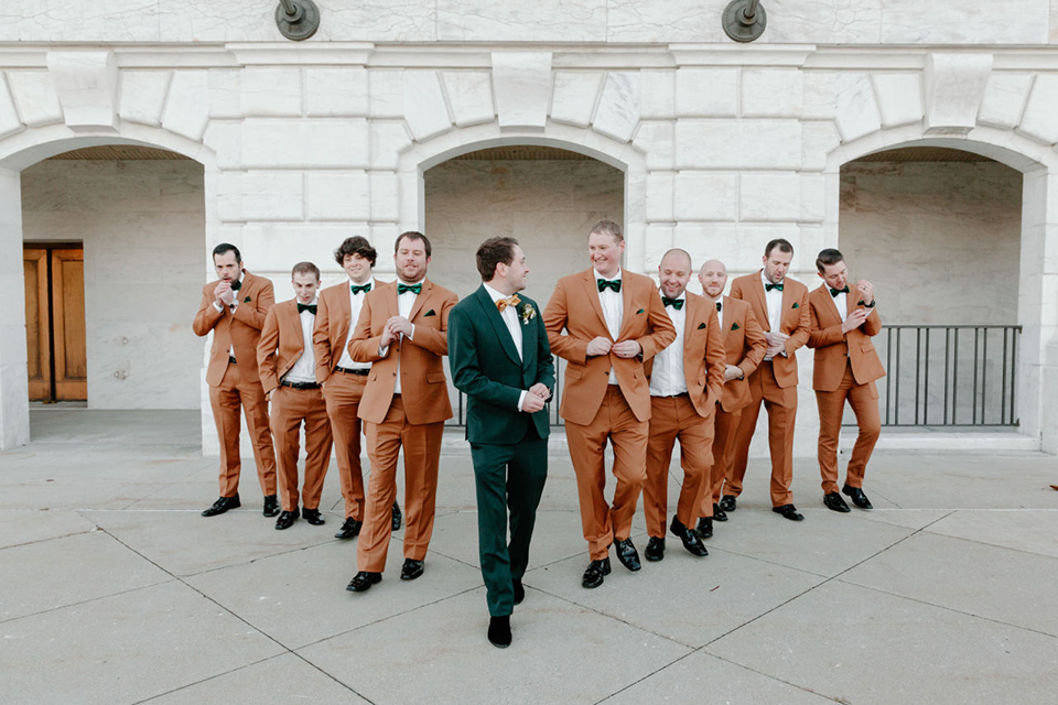  bride in a trumpet style gown and the groom in a green suit, the groomsmen in caramel notch lapel suits and the bridesmaids in ivory gowns 